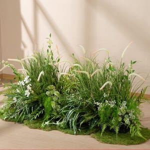 Puede incluir: Un exuberante arreglo de vegetación artificial, con altas hierbas verdes con cabezas de semillas plumosas, varias tonalidades de follaje verde y pequeñas flores blancas. La exhibición está sobre un lecho de musgo, creando un acento decorativo natural.