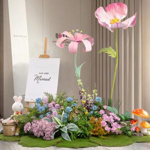 Pode incluir: Uma exibição de casamento extravagante com grandes flores de papel rosa, uma placa que diz "we are Married" e uma variedade de flores e vegetação artificiais coloridas. Uma pequena figura de coelho e decorações de cogumelos adicionam ao tema de conto de fadas.