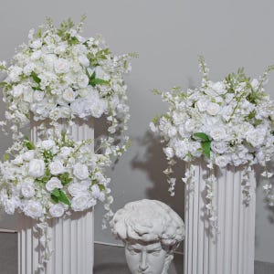 Puede incluir: Arreglos florales blancos sobre columnas estriadas, con rosas y vegetación colgante. Un busto clásico está frente a las columnas. Las flores son una mezcla de rosas blancas y otras flores blancas.