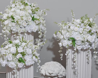 May include: White floral arrangements on fluted columns, with roses and trailing greenery. A classical bust is in front of the columns. The flowers are a mix of white roses and other white blossoms.