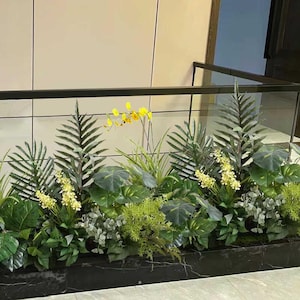 May include: A lush arrangement of artificial plants, including large green leaves, ferns, and yellow orchids, fills a rectangular planter. The planter is made of black marble and sits beneath a black metal railing.