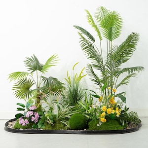 May include: An artificial tropical garden arrangement with a variety of green plants, including palm fronds, grasses, and flowering plants. The arrangement has a moss-covered base with small stones and a black oval border.