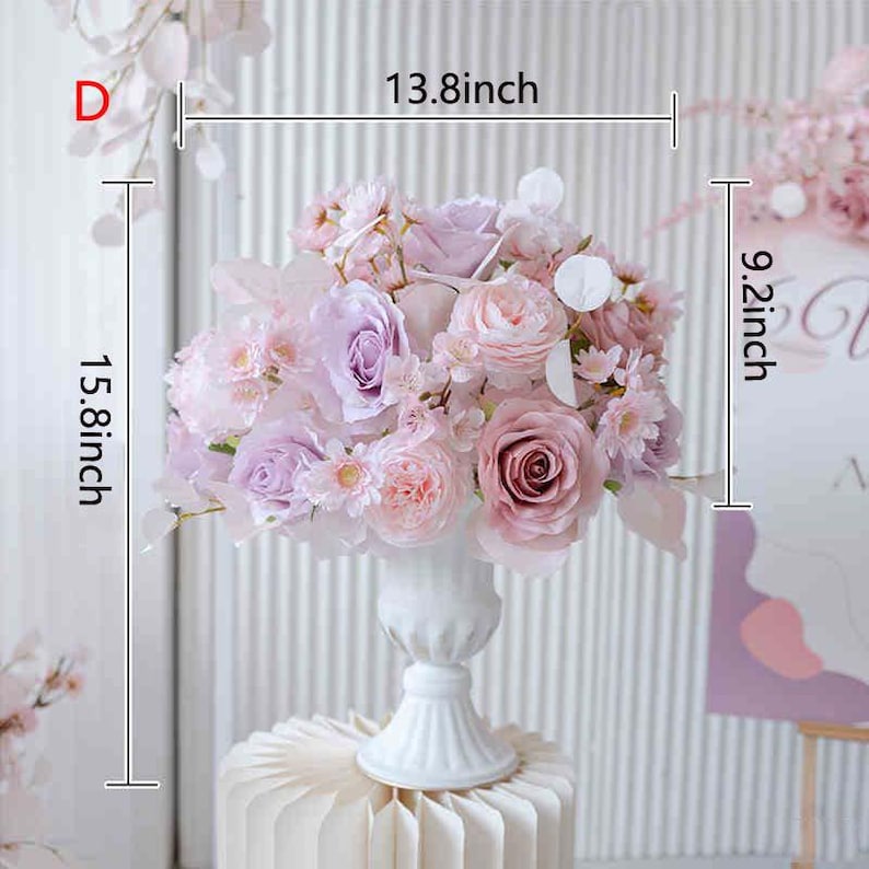 May include: A floral arrangement featuring a variety of pink and purple flowers, including roses and smaller blossoms, in a white vase. The arrangement measures approximately 15.8 inches high, 13.8 inches wide, and 9.2 inches deep.