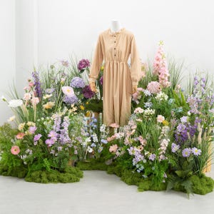 Könnte beinhalten: Ein beigefarbenes Kleid mit langen Ärmeln und Knöpfen an einer Schaufensterpuppe. Das Kleid ist von einer lebendigen Ausstellung farbenfroher Blumen und Grünpflanzen umgeben, die eine natürliche und elegante Umgebung schaffen.