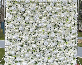 May include: A large, square backdrop densely covered with white artificial flowers, including roses and hydrangeas. The flowers are arranged to create a full, textured surface, ideal for event decorations. The backdrop is supported by a metal frame.