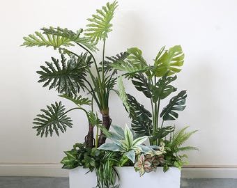 May include: A lush arrangement of artificial tropical plants in a white rectangular planter. The arrangement features large, green, split-leaf philodendron and monstera leaves, with smaller ferns and other foliage. The plants have a realistic appearance.