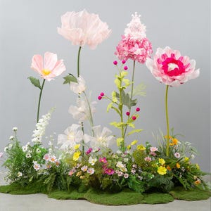 Puede incluir: Un vibrante arreglo floral con grandes flores de papel en tonos rosa y blanco, con centros amarillos. El arreglo incluye una variedad de flores más pequeñas y vegetación, sobre un fondo gris. La base está cubierta de musgo verde.
