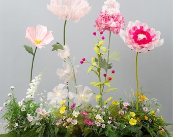 May include: A vibrant floral arrangement featuring large, paper-like flowers in shades of pink and white, with yellow centers. The arrangement includes a variety of smaller flowers and greenery, set against a gray backdrop. The base is covered in green moss.