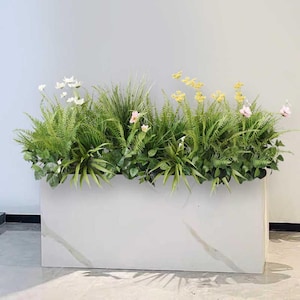 Peut inclure: Un bac à fleurs rectangulaire blanc avec un motif marbré, rempli d'un assortiment de plantes vertes et de fleurs colorées. L'arrangement comprend des fougères, des graminées et de petites fleurs roses, jaunes et blanches, créant un affichage vibrant.