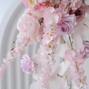 May include: A close-up of a floral arrangement featuring various shades of pink and white flowers. The arrangement includes roses, peonies, and cascading blossoms, creating a soft and romantic aesthetic. The flowers are set against a white background.