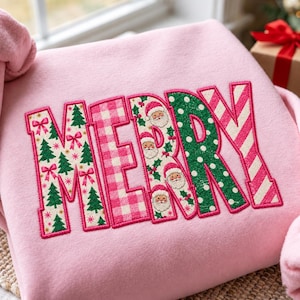 May include: A light pink sweatshirt featuring the word MERRY in large, block-style letters. Each letter has a unique pattern: green Christmas trees and pink bows, pink gingham, Santa Claus faces with holly, green polka dots, and pink diagonal stripes.