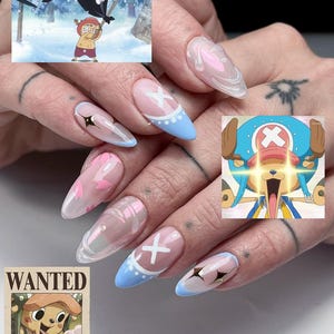 May include: Almond-shaped nail art with a One Piece theme. The nails feature pink and blue designs, white crosses, and gold star details. A "Wanted" poster of Tony Tony Chopper is also visible in the image.