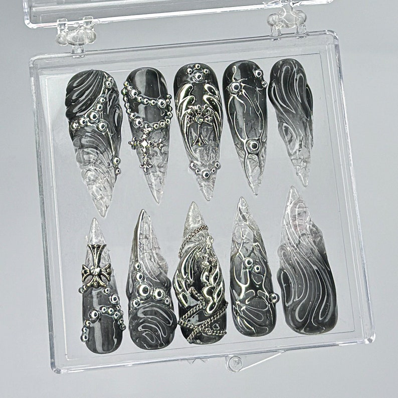 Puede incluir: Una caja de pl&aacute;stico transparente con 10 dise&ntilde;os de arte de u&ntilde;as negros y plateados. Los dise&ntilde;os presentan patrones intrincados, remolinos y adornos.