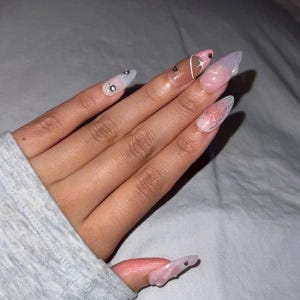 May include: Close-up of a hand with long, almond-shaped nails. The nails are painted with a white base and have different designs, including a heart, a star, and a butterfly. Some nails have small rhinestones.