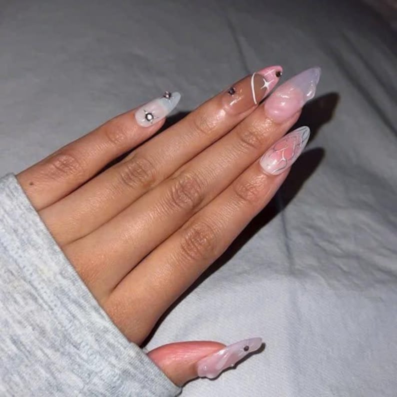 May include: Close-up of a hand with long, almond-shaped nails. The nails are painted with a white base and have different designs, including a heart, a star, and a butterfly. Some nails have small rhinestones.