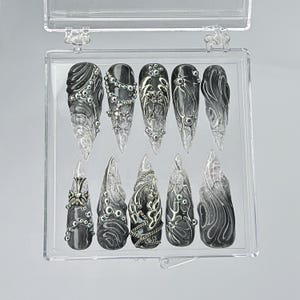 Puede incluir: Una caja de pl&aacute;stico transparente con 10 u&ntilde;as postizas. Las u&ntilde;as son negras con detalles y dise&ntilde;os plateados. Las u&ntilde;as est&aacute;n dispuestas en dos filas de cinco.