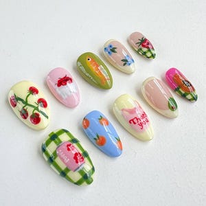 Peut inclure: Ensemble de dix faux ongles avec diff&eacute;rents motifs, dont un poisson, des fraises, une canette de soda, des oranges et les mots "Merci".