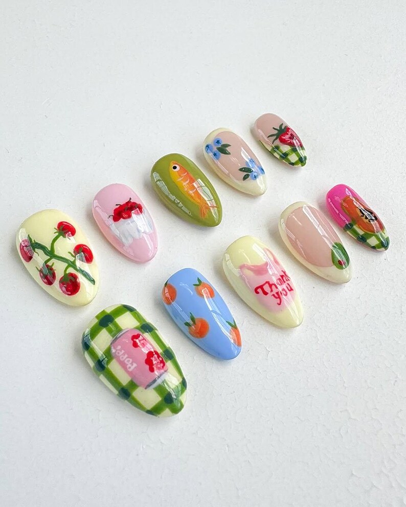 Peut inclure: Ensemble de dix faux ongles avec diff&eacute;rents motifs, dont un poisson, des fraises, une canette de soda, des oranges et les mots "Merci".