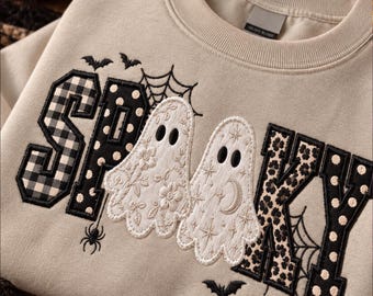 May include: A beige crewneck sweatshirt featuring embroidered applique lettering that spells SPOOKY. The letters incorporate black and white buffalo plaid, polka dot, and floral patterns. Two white ghost figures are centered, surrounded by spiderwebs, bats, and a small spider.