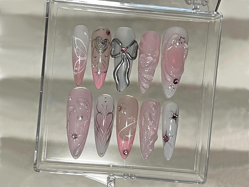 May include: A set of ten false nails with different designs. The nails are all a light pink color with white and silver accents. Some of the designs include butterflies, hearts, and bows.