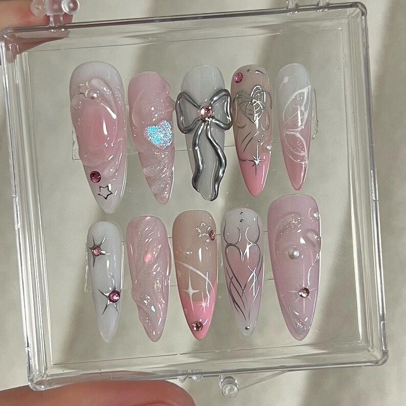 May include: A set of ten pink and white artificial nails with various designs, including a bow, a butterfly, and swirls. The nails are displayed in a clear plastic box.