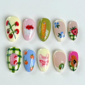 Peut inclure: Ensemble de dix faux ongles avec diff&eacute;rents motifs, dont une plante de tomates, un bol de cerises, un poisson, des myrtilles, une fraise, un motif &agrave; carreaux, des p&ecirc;ches, un message de remerciement, une feuille verte et une papaye.