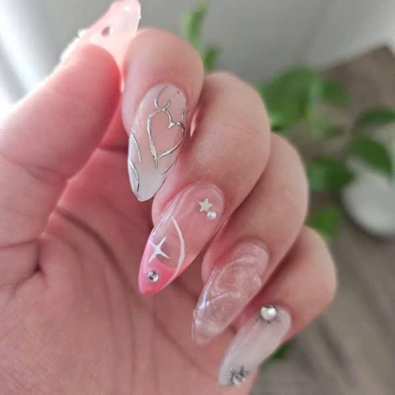 May include: Close-up of a hand with long, oval-shaped nails painted with a pink and white ombre design. The nails are decorated with silver glitter, stars, and hearts.