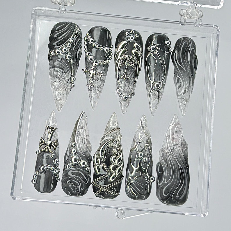 Puede incluir: Un conjunto de diez u&ntilde;as postizas negras y plateadas con dise&ntilde;os intrincados. Las u&ntilde;as presentan una variedad de patrones, incluyendo espirales, cadenas y cruces. Las u&ntilde;as se muestran en una caja de pl&aacute;stico transparente.