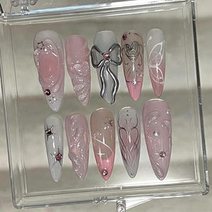 May include: A set of ten pink and white false nails with various designs, including a bow, a butterfly, and a heart. The nails are displayed in a clear plastic case.