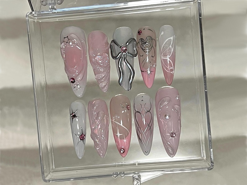 May include: A set of ten pink and white false nails with various designs, including a bow, a butterfly, and a heart. The nails are displayed in a clear plastic case.