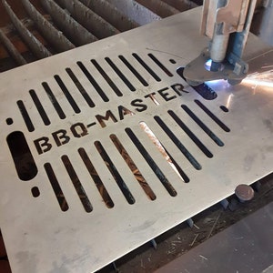 K&ouml;nnte beinhalten: Eine Metallplatte mit einem Gittermuster und den ausgeschnittenen Worten "BBQ-MASTER". Ein Plasmaschneider schneidet die Metallplatte.