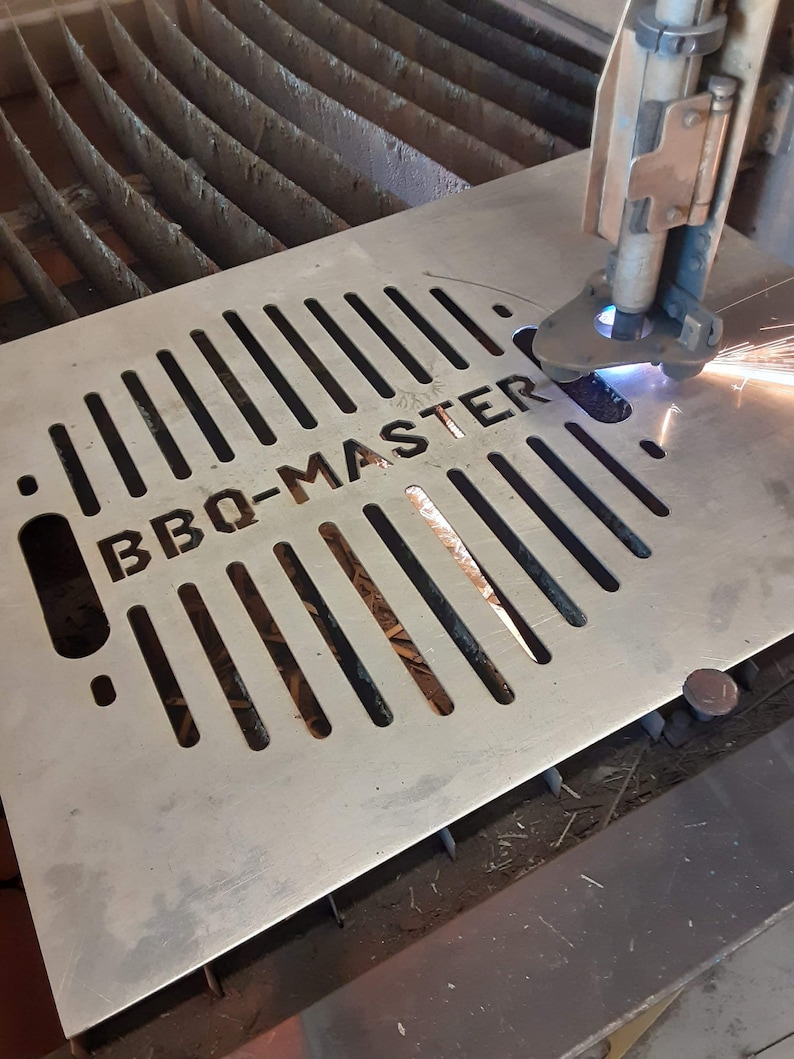 K&ouml;nnte beinhalten: Eine Metallplatte mit einem Gittermuster und den ausgeschnittenen Worten "BBQ-MASTER". Ein Plasmaschneider schneidet die Metallplatte.