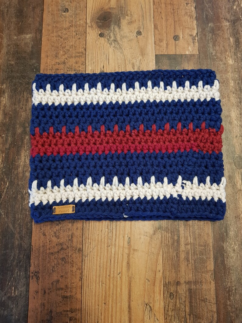 Pu&ograve; includere: Un scaldacollo lavorato a uncinetto con strisce blu navy, bianco e bordeaux. Lo scaldacollo ha un piccolo cartellino in pelle marrone con la parola "Needle" su di esso.