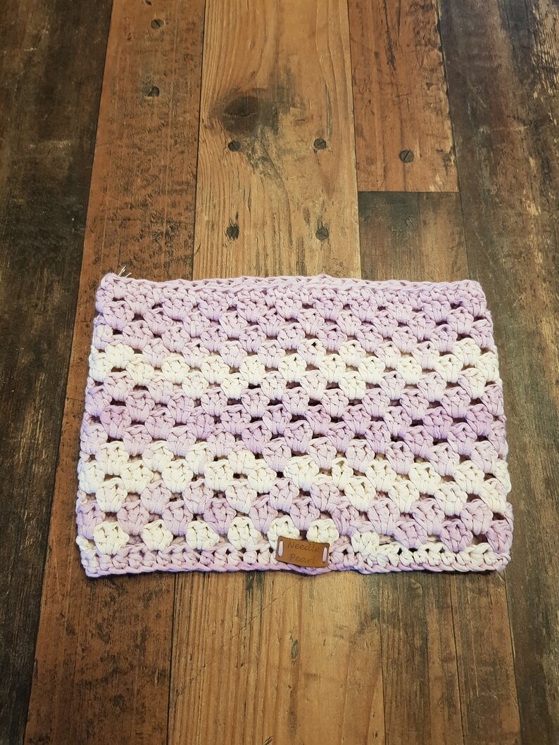 Pu&ograve; includere: Una coperta all'uncinetto con un motivo a strisce viola e bianche. La coperta ha un piccolo cartellino in pelle marrone con la parola "Needle" su di esso.