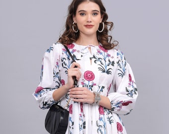 May include: White dress with a floral print featuring pink flowers and blue leaves. The dress has long sleeves, a round neckline, and a knee-length skirt. A black purse is worn over the shoulder.
