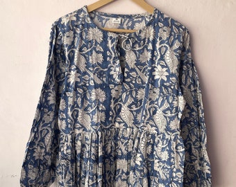 May include: A long-sleeved midi dress featuring a blue and white floral and bird block print pattern. The garment has a round neckline with a small front slit and a tie closure, a gathered waistline, and long sleeves with elasticized cuffs.