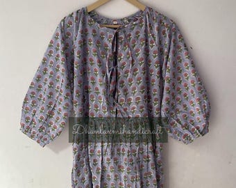 May include: A light purple knee-length dress featuring a repeating pattern of small pink flowers with green stems. The dress has three-quarter length puff sleeves, a round neckline with a central tie closure, and a gathered waistline.
