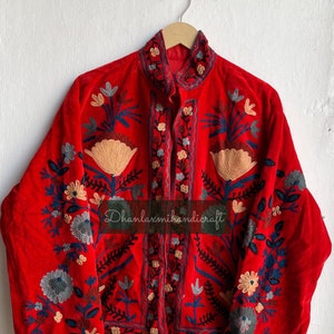 May include: A red velvet jacket with intricate floral embroidery in shades of blue, green, and cream. The jacket has a mandarin collar and a button closure. The text "Dhanlaxmihandicraft" is visible on the front of the jacket.