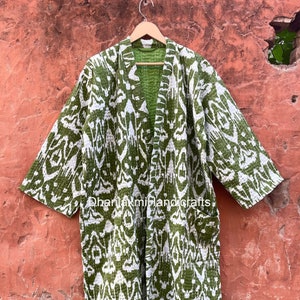 May include: A green and white patterned kimono-style robe hanging on a wooden hanger. The robe features a repeating ikat design. The background is a textured, reddish-brown wall. The robe has pockets and a green lining.