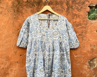 May include: A short-sleeved, light blue and white floral print dress. The dress features a V-neckline and a tiered skirt. The fabric is patterned with stylized flowers and leaves. The dress is hanging on a wooden hanger.