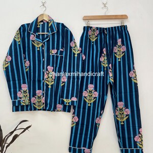 Puede incluir: Un conjunto de pijama de dos piezas con un patrón de rayas azul marino y azul claro. El conjunto incluye una camisa con botones y pantalones, ambos adornados con motivos florales rosas. Ribetes blancos acentúan el cuello, los puños y los bolsillos. El pijama está colgado.