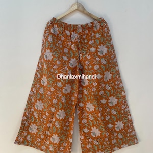May include: Wide-leg pants in a burnt orange hue, adorned with a floral pattern in white and light green. The pants are hanging on a wooden hanger against a plain white background. The text "Dhanlaxmihandi" is visible.