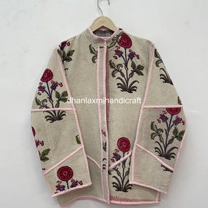 May include: A beige quilted jacket featuring a floral pattern with pink flowers and green stems. The jacket has a mandarin collar, long sleeves, and light pink piping along the seams and edges. The text dhanlaxmihandicraft is visible.