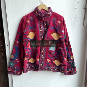 May include: A red velvet jacket with intricate floral embroidery in shades of blue, yellow, and green. The jacket has a mandarin collar and a button closure. The text "Dhanlaxmihandicraft" is visible on the front of the jacket.