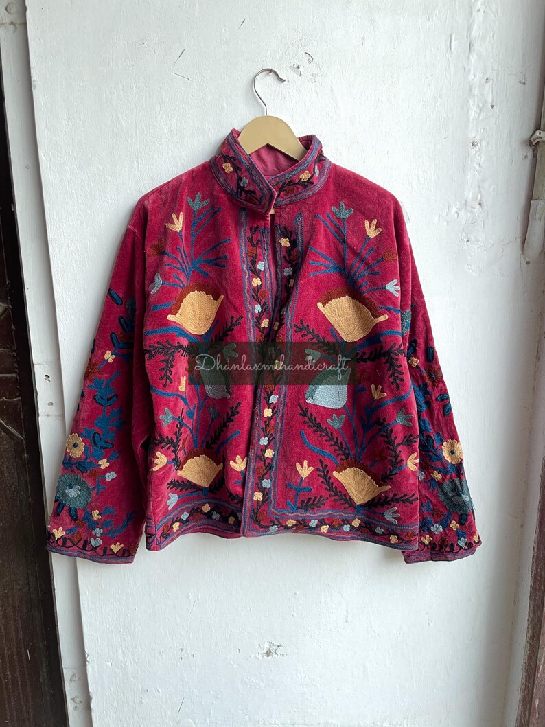 May include: A red velvet jacket with intricate floral embroidery in shades of blue, yellow, and green. The jacket has a mandarin collar and a button closure. The text "Dhanlaxmihandicraft" is visible on the front of the jacket.