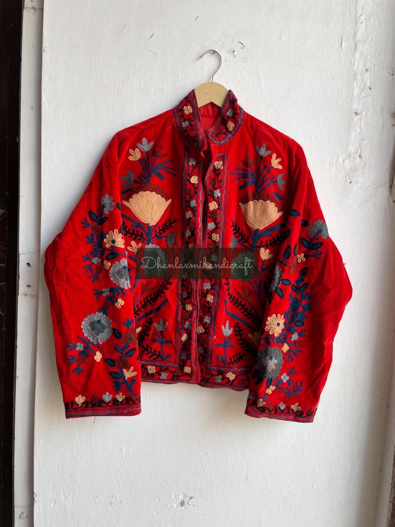 May include: A red velvet jacket with intricate floral embroidery in shades of blue, green, and cream. The jacket has a mandarin collar and a button closure. The text "Dhanlaxmihandicraft" is visible on the front of the jacket.