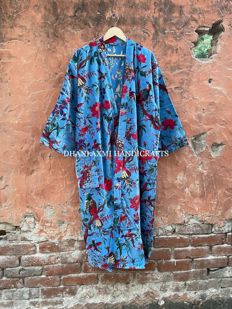 May include: A blue cotton robe with a floral and bird print. The robe has a long, flowing silhouette and a tie closure at the front. The text "DHANLAXMI HANDICRAFTS" is printed on the bottom of the robe.