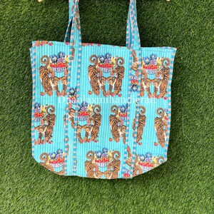 以下が含まれることがあります： キルティング加工が施されたターコイズブルーのトートバッグ。果物の入ったボウルを持ったトラが繰り返し描かれたパターン。バッグには長いストラップが2本付いている。