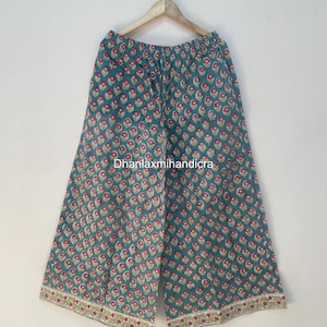 Puede incluir: Pantalones palazzo de pierna ancha en color verde azulado con un estampado floral repetido en rojo y blanco. Los pantalones están colgados de una percha de madera. El dobladillo inferior presenta un borde decorativo con un diseño floral.