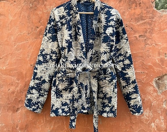 Könnte beinhalten: Eine marineblaue gesteppte Jacke mit einem weißen Blumenmuster und einem passenden Bindegürtel. Die Jacke hängt an einem Holzbügel an einer Backsteinwand.
